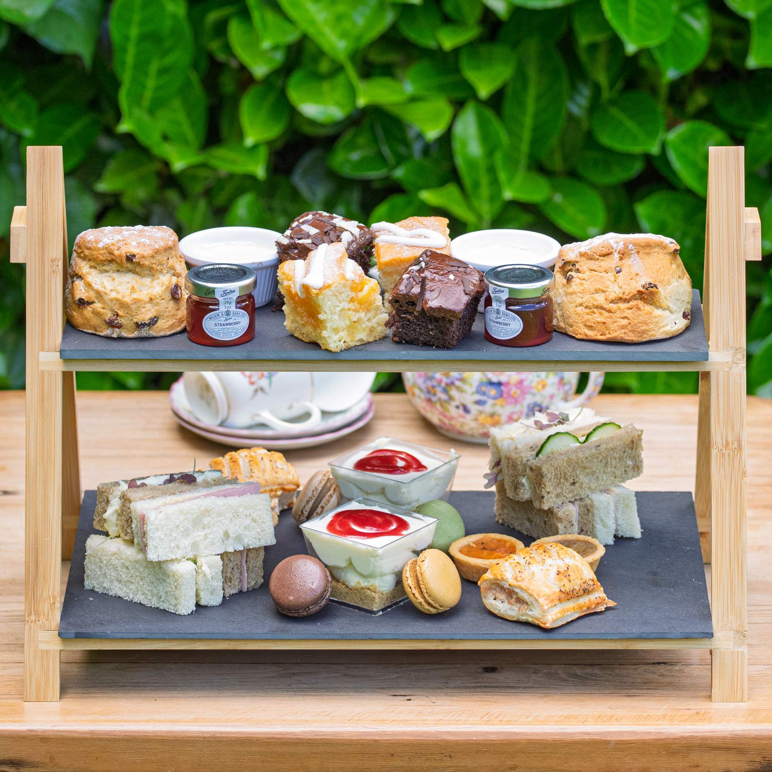 Barnsdale Afternoon Tea from The Helenium Tea Room at Barnsdale Gardens. Two-tiered serving tray with assorted finger foods on a wooden table outdoors.