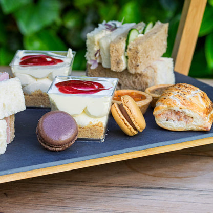 Barnsdale Afternoon Tea from The Helenium Tea Room at Barnsdale Gardens. Assorted finger foods including sandwiches, pastries, and small dishes on a slate board with a green leafy background.