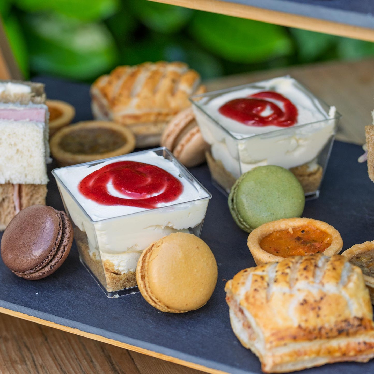 Barnsdale Afternoon Tea from The Helenium Tea Room at Barnsdale Gardens. Assorted pastries including tarts, macarons, and pies on a slate surface with a blurred green background.