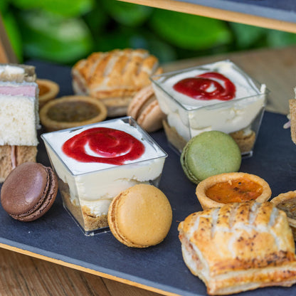Barnsdale Afternoon Tea from The Helenium Tea Room at Barnsdale Gardens. Assorted pastries including tarts, macarons, and pies on a slate surface with a blurred green background.