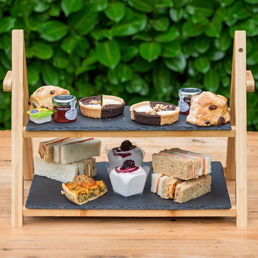 Vegan Afternoon Tea from the Helenium Tea Room at Barnsdale Gardens. A two-tier wooden serving stand holding assorted sandwiches, scones, pastries, and small dessert cakes, photographed on a wooden table with a leafy green hedge in the background.