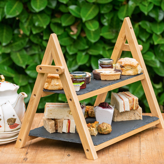 Vegan Afternoon Tea from the Helenium Tea Room at Barnsdale Gardens. A two-tier wooden serving stand with dark slate shelves, styled with assorted sandwiches, scones, pastries, and small jars of preserves on a wooden table in front of a green leafy background.