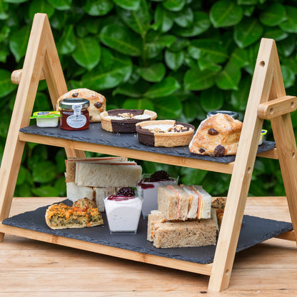 Vegan Afternoon Tea from the Helenium Tea Room at Barnsdale Gardens.. A two-tier wooden A-frame serving tray with dark slate trays, styled with assorted sandwiches, scones, cakes, and small desserts, set against a background of green foliage.
