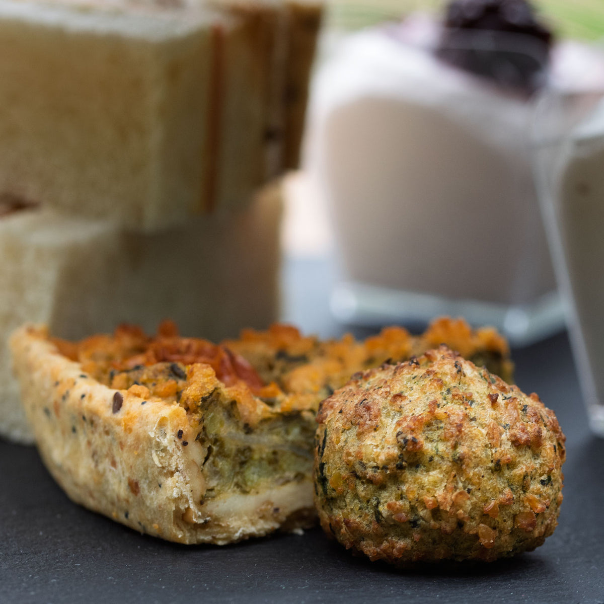 Vegan Afternoon Tea from the Helenium Tea Room at Barnsdale Gardens. A close-up food photograph showing a vegan falafel and vegan mini-quiche on a dark slate plate, with blurred sandwiches and vegan Eton mess puddings in the background.