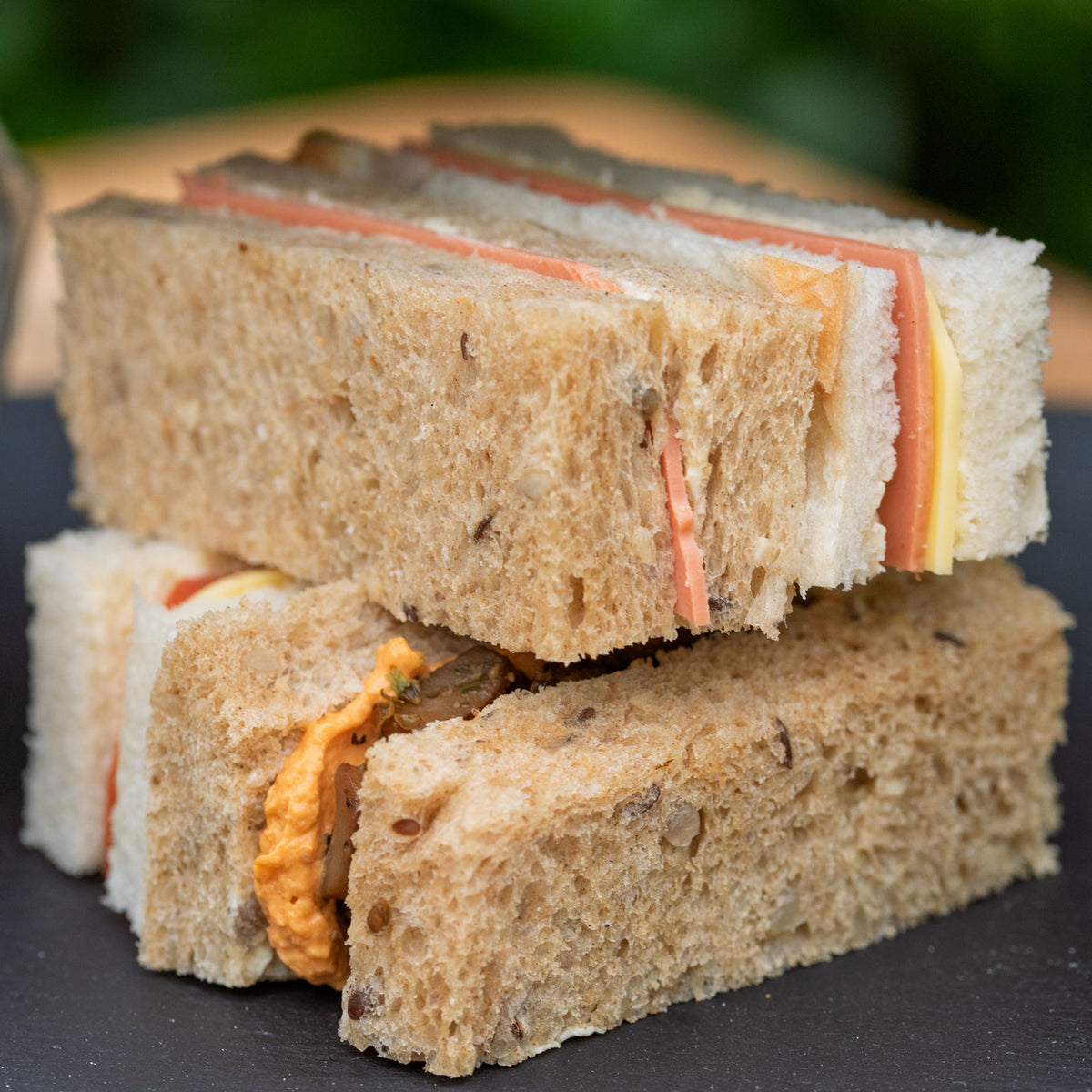 Vegan Afternoon Tea from the Helenium Tea Room at Barnsdale Gardens. Stacked vegan fnger sandwiches on a dark slate surface with a blurred green outdoor background. 