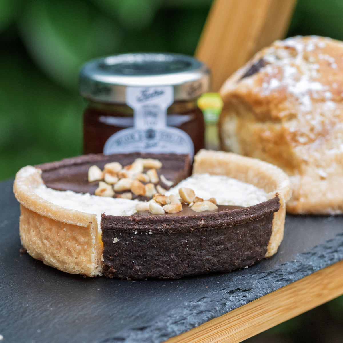 Vegan Afternoon Tea from the Helenium Tea Room at Barnsdale Gardens. A close-up of a chocolate tart with a cream-colored center and chopped nuts on top, served on a dark slate board with a jar of preserve and a vegan fruit scone in the background.
