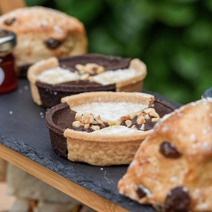 Vegan Afternoon Tea from the Helenium Tea Room at Barnsdale Gardens.. Chocolate tart and scones arranged on a dark slate serving board, with a blurred green outdoor background.