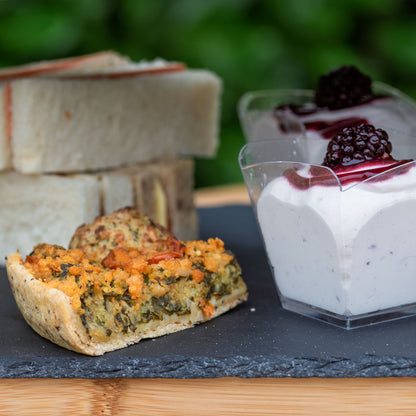 Vegan Afternoon Tea from the Helenium Tea Room at Barnsdale Gardens.. A close-up food scene showing a slice of vegan quiche in the foreground, stacked vegan finger sandwiches on the left and two clear cups of vegan Eton mess topped with blackberries on the right, arranged on a dark slate board over a wooden surface with a blurred green outdoor background.