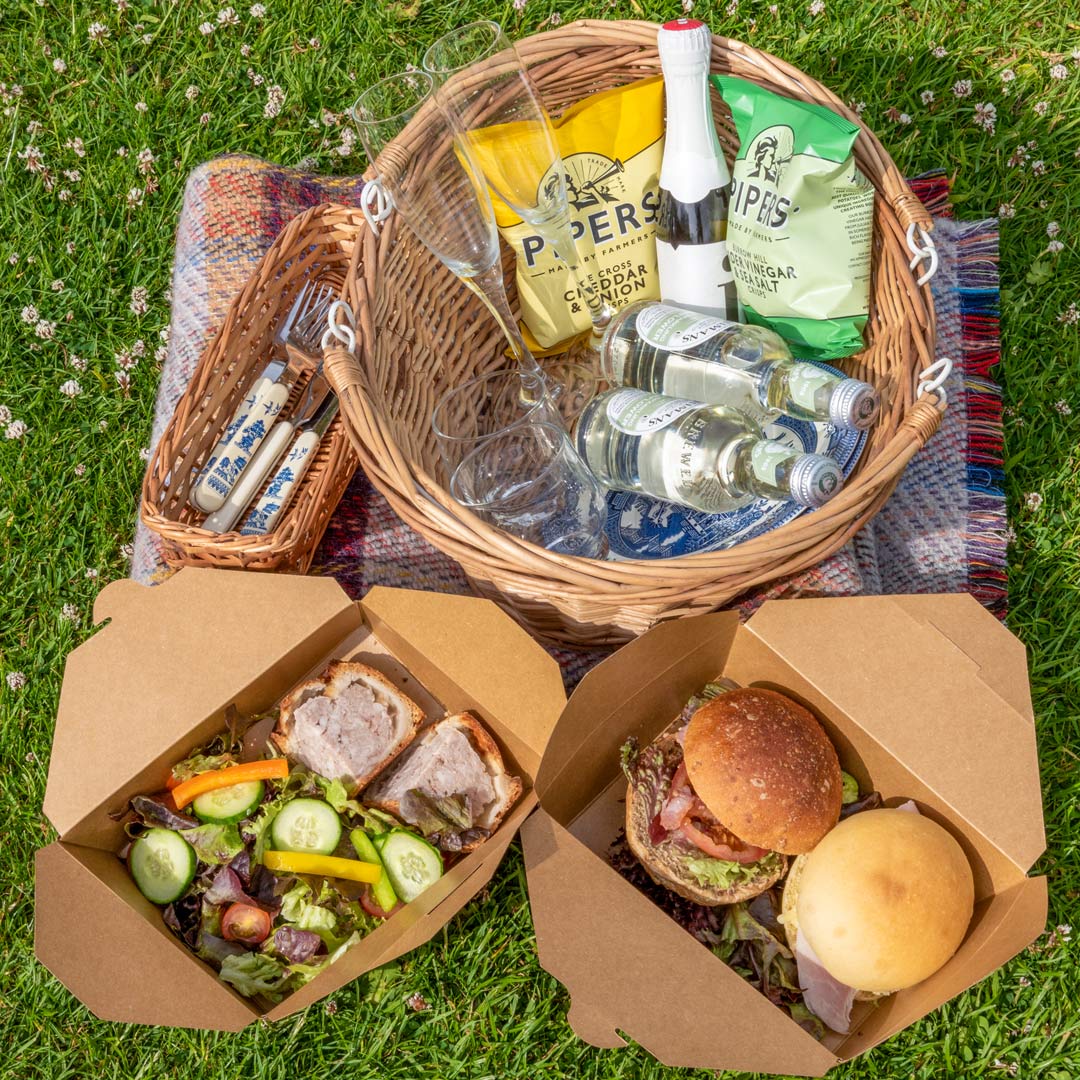 Overhead image of the picnic basket for two, on a picnic blanket. Comprising: two packets of crisps; two soft drinks, with glasses; filled rolls; mixed salad; two portions of pork pie. Small basket of Victorian style cutlery.